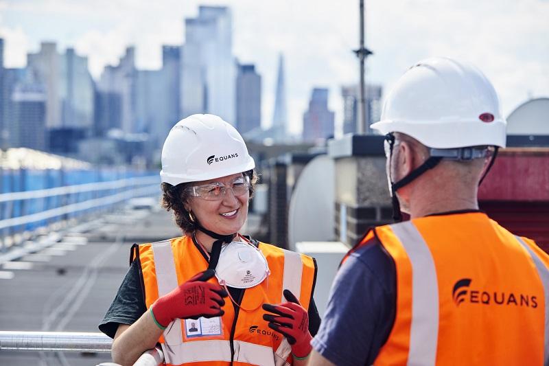 Two Equans employees stood on a rooftop wearing hard hats with chin straps